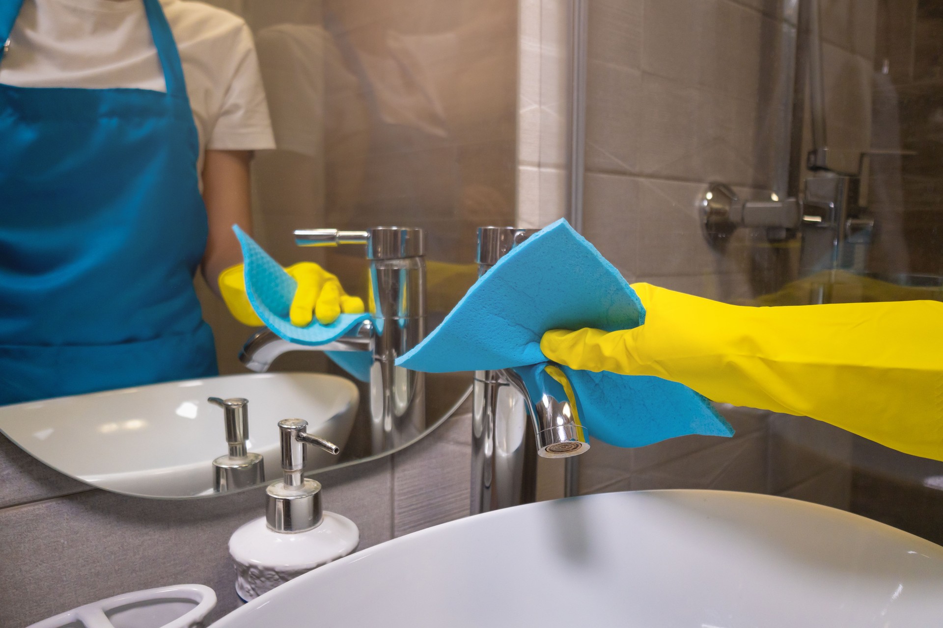 Close-up of a woman 's hand in yellow rubber gloves wiping the faucet with a blue cleaning cloth in a modern bathroom. Concept of professional cleaning
